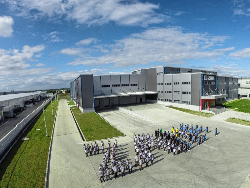 Menschen bilden den Schriftzug ZAMA, im Hintergrund ein Werk.