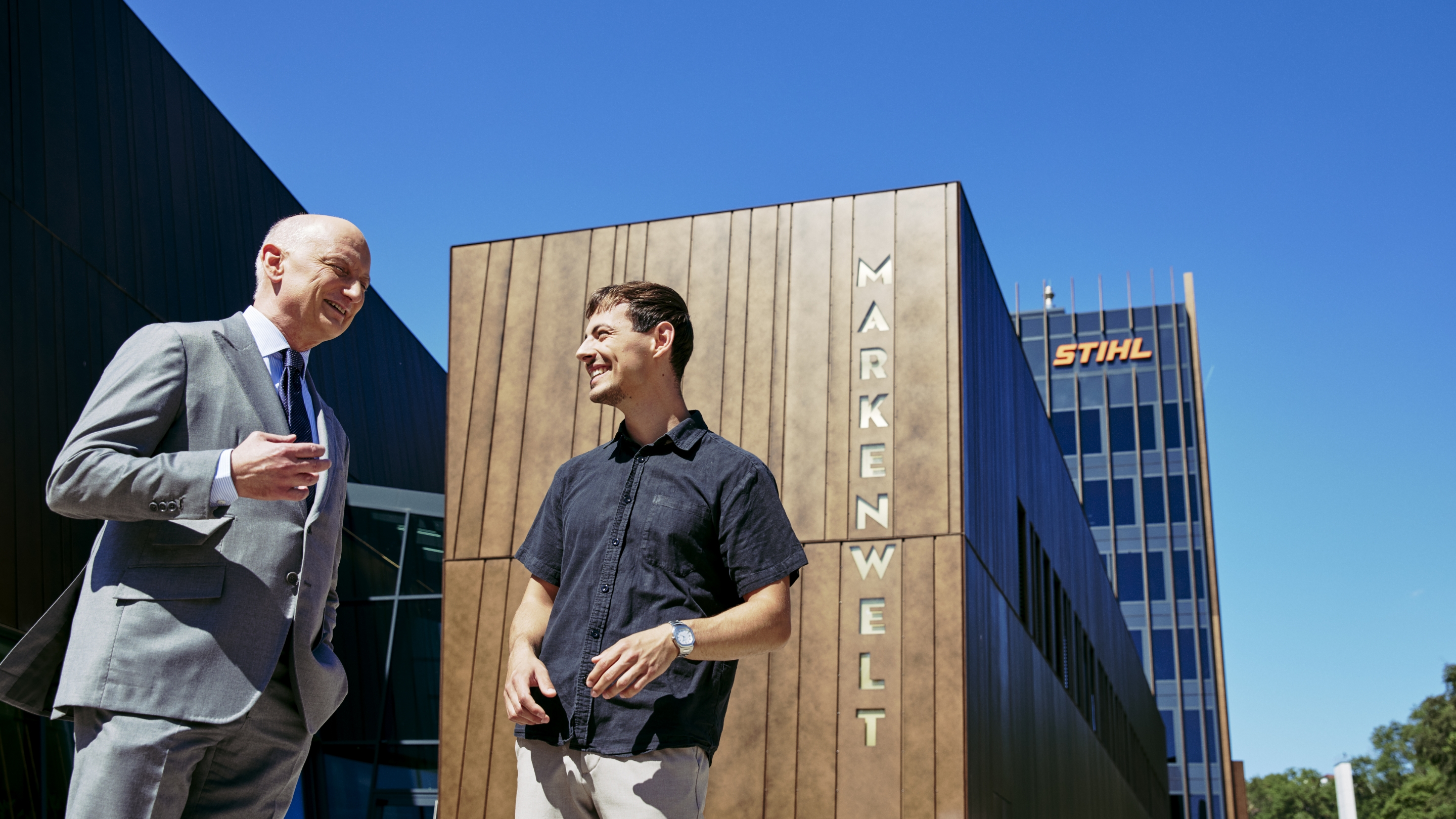 Dr. Nikolas Stihl und Tamino Soubawi unterhalten sich vor einem modernen Geb&auml;ude mit den Schriftz&uuml;gen &bdquo;Markenwelt&ldquo; und &bdquo;STIHL&ldquo;.