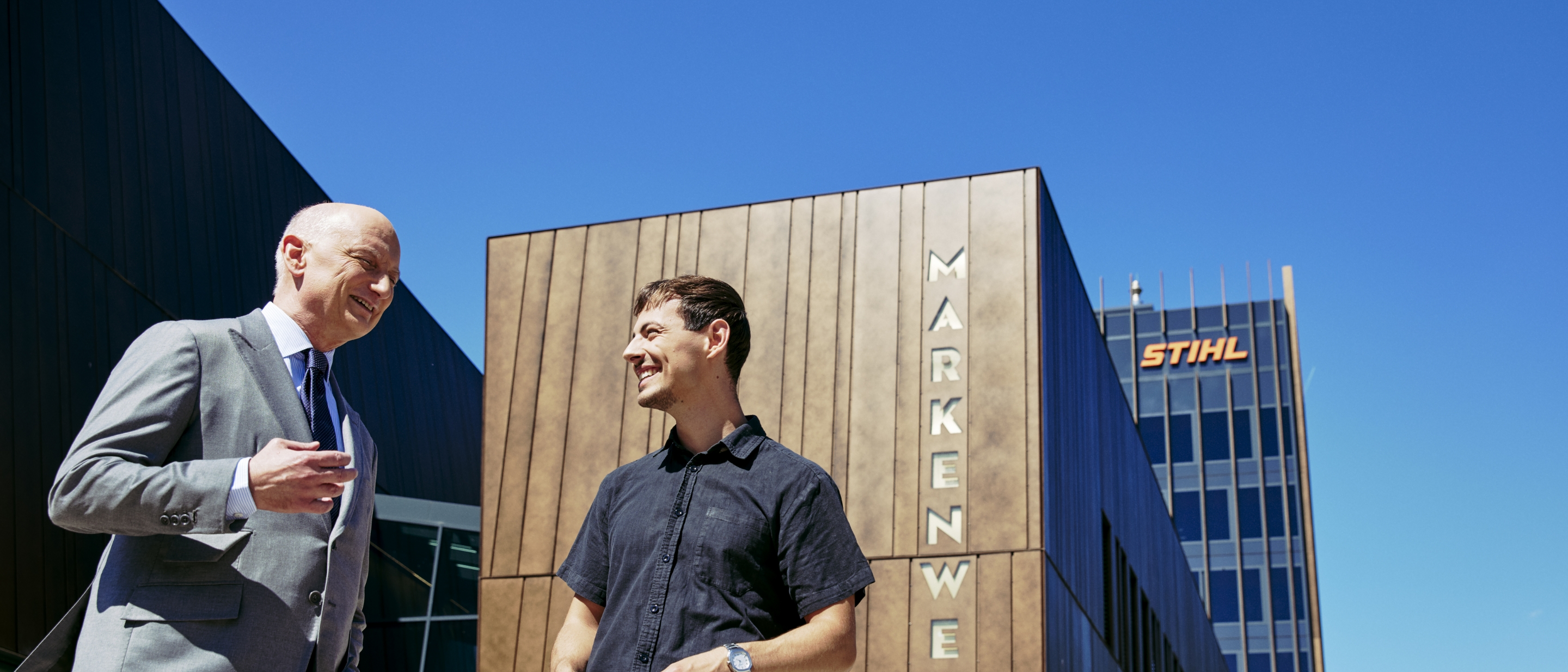 Dr. Nikolas Stihl und Tamino Soubawi unterhalten sich vor einem modernen Geb&auml;ude mit den Schriftz&uuml;gen &bdquo;Markenwelt&ldquo; und &bdquo;STIHL&ldquo;.