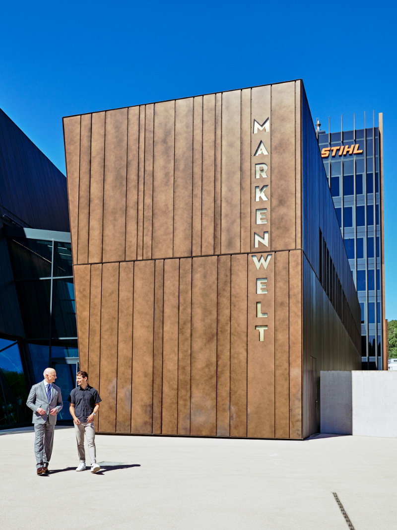 Links vor einem braunen Geb&auml;ude mit der Aufschrift &bdquo;Markenwelt&ldquo; gehen zwei M&auml;nner.