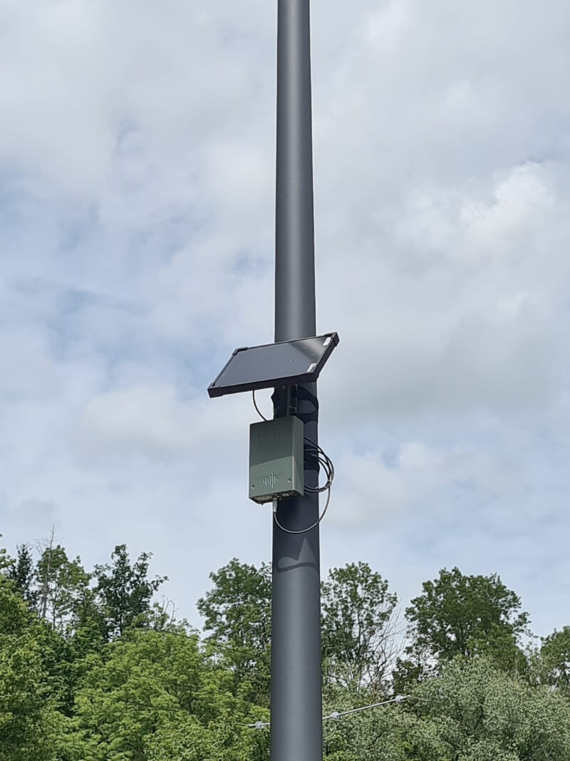 Ein Laternenpfahl, an dem ein rechteckiges K&auml;stchen und eine Solarzelle angebracht sind, rundherum B&auml;ume.